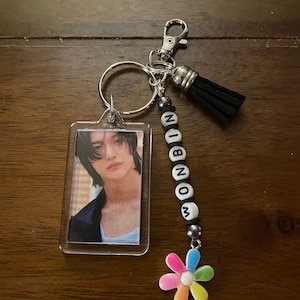 May include: A keychain with a clear plastic photo frame featuring a man with dark hair. The keychain also has a black tassel, a silver charm, and a beaded charm with the word "WONBIN" spelled out in white letters.