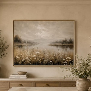 May include: A rectangular landscape painting in a thin gold frame depicting a serene lake surrounded by rolling hills and a field of white wildflowers in muted earth tones. The artwork is displayed on a beige wall above a kitchen counter.