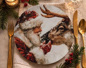 May include: A round ceramic dinner plate featuring a detailed illustration of Santa Claus wearing a red hat and coat, touching noses with a reindeer. The scene includes white snowflakes against a light background, creating a festive winter atmosphere.