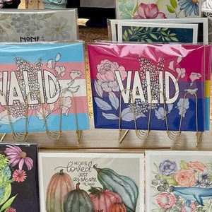 May include: Four framed art prints with floral designs and the word "VALID" in bold lettering. The prints are displayed on a gold metal stand. The prints are on a wooden surface.