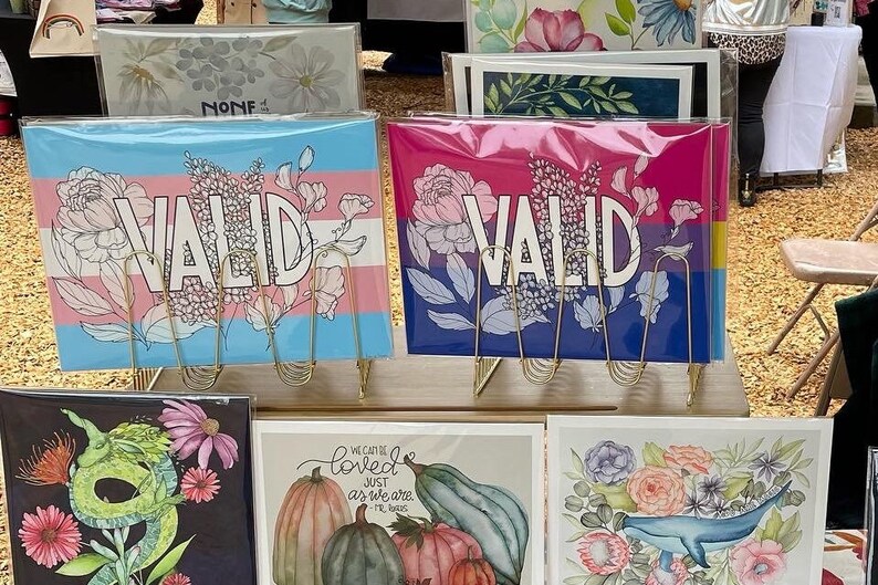 May include: Four framed art prints with floral designs and the word "VALID" in bold lettering. The prints are displayed on a gold metal stand. The prints are on a wooden surface.