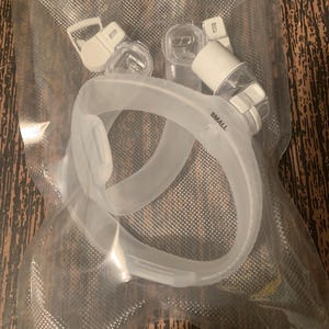 May include: A clear plastic nasal mask for sleep apnea with a white connector and the word "SMALL" printed on it.