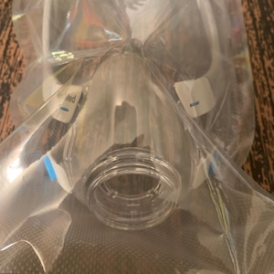 May include: A clear, plastic medical mask, sealed in a transparent bag. The mask has a circular opening and is labeled with the word "Med". The mask is designed for medical use.