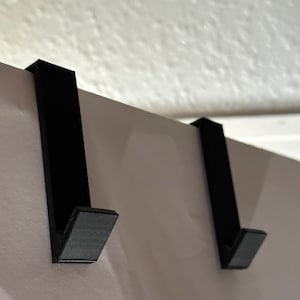 Può includere: Due ganci neri sopra la porta. I ganci sono realizzati in un solido materiale nero opaco e sono progettati per essere appesi sopra la parte superiore di una porta. I ganci hanno un design semplice e minimalista, con una sezione verticale diritta e un gancio ad angolo retto.