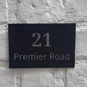 Puede incluir: Un letrero rectangular negro con el número de casa "21" y las palabras "Premier Road" en un color más claro. El letrero está montado en una pared de ladrillos blancos.