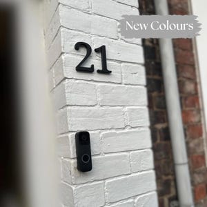 Peut inclure: Numéro de maison noir "21" fixé sur un mur de briques blanches, avec une sonnette noire en dessous. Le texte "New Colours" est au-dessus des chiffres. Le mur fait partie de l'extérieur d'un bâtiment.