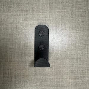 May include: A black wall hook with a rounded top and a curved base. Two textured, circular pads are attached to the hook's vertical section. The hook is designed for hanging items and is made of a solid, matte material.