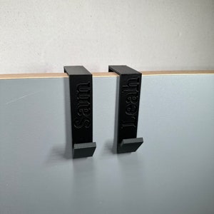 Könnte beinhalten: Zwei schwarze Türhaken mit den eingravierten Namen "Sam" und "Leah". Sie hängen an einer grauen Oberfläche mit einer Holzkante.