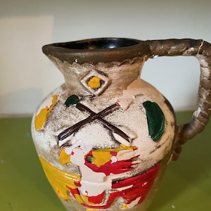Peut inclure: Un pichet en céramique blanche avec une anse brune. Le pichet est décoré d'un motif coloré d'un oiseau, de formes géométriques et d'une croix. Le pichet a un aspect rustique et vintage.