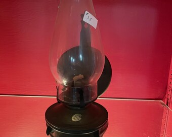 May include: A black metal oil lamp with a clear glass chimney. The lamp has a round base and a decorative label on the side. The lamp is on a red shelf.