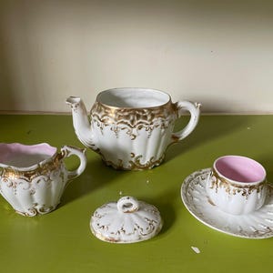 Può includere: Un set da tè bianco con dettagli dorati. Il set comprende una teiera, un creamer, una zuccheriera e un piattino. La teiera ha un corpo rotondo e un beccuccio laterale. Il creamer e la zuccheriera hanno un corpo rotondo e un manico laterale. Il piattino è rotondo e ha un bordo rialzato.