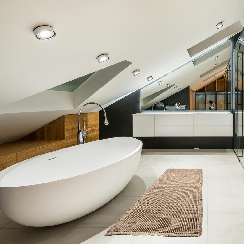 Op de afbeelding: Een vrijstaand wit bad met een chromen kraan in een moderne badkamer met een witte wastafel en een bruin vloerkleed.