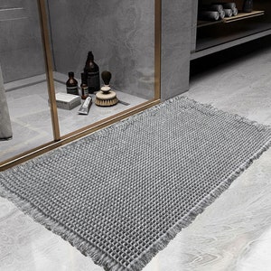 Op de afbeelding: Een grijze badmat met een getextureerd wafelidee en franjes. De mat ligt op een grijze tegelvloer in een badkamer.