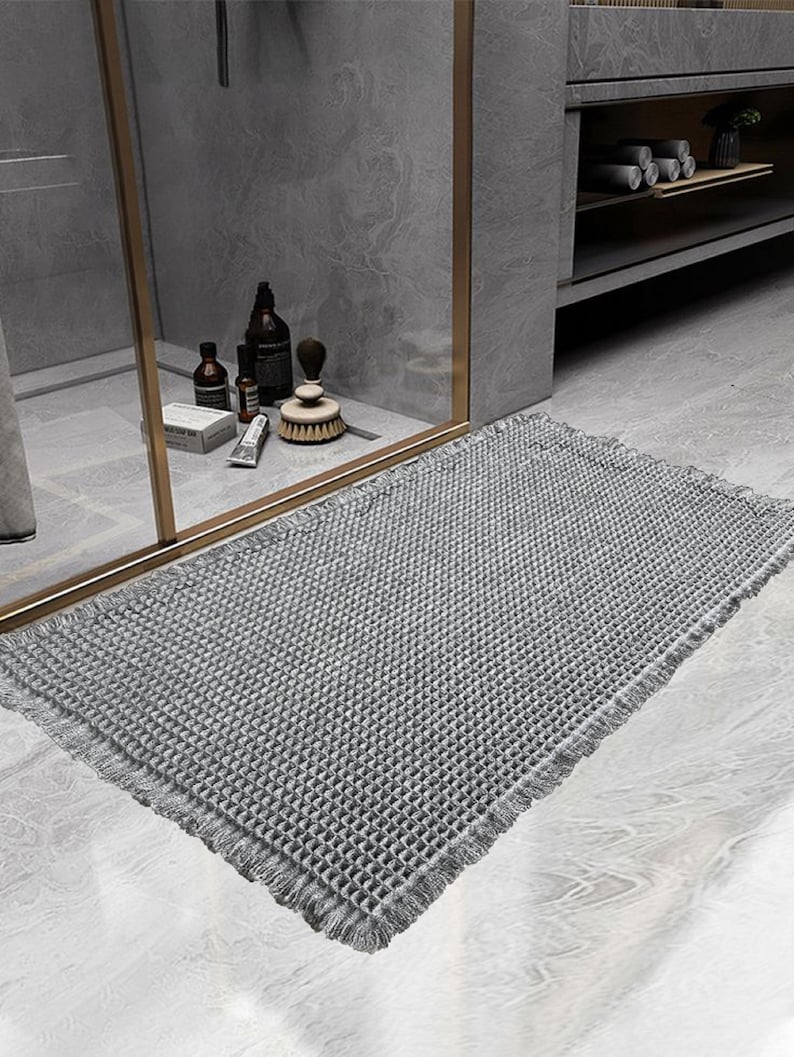 Op de afbeelding: Een grijze badmat met een getextureerd wafelidee en franjes. De mat ligt op een grijze tegelvloer in een badkamer.