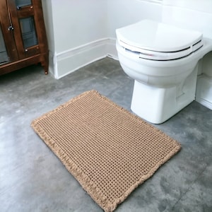 Op de afbeelding: Een bruin geweven badkamerkleed met franjes aan de randen. Het kleed ligt voor een wit toilet in een badkamer.