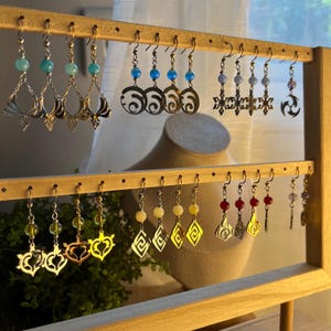 May include: A wooden earring display stand with a variety of dangle earrings. The earrings have diverse designs, including floral, geometric, and abstract shapes, with accents of blue, green, red, and yellow beads. The earrings are made of metal.
