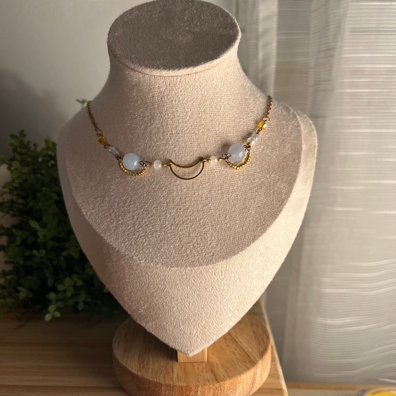 Op de afbeelding: Een delicate gouden ketting met een halve maan bedel en blauwe en witte kralen.