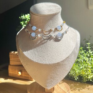 Op de afbeelding: Twee delicate kettingen met halve maan hangers en lichtblauwe kralen op een witte mannequinbuste. De kettingen zijn gelaagd en hebben zilveren en gouden accenten.