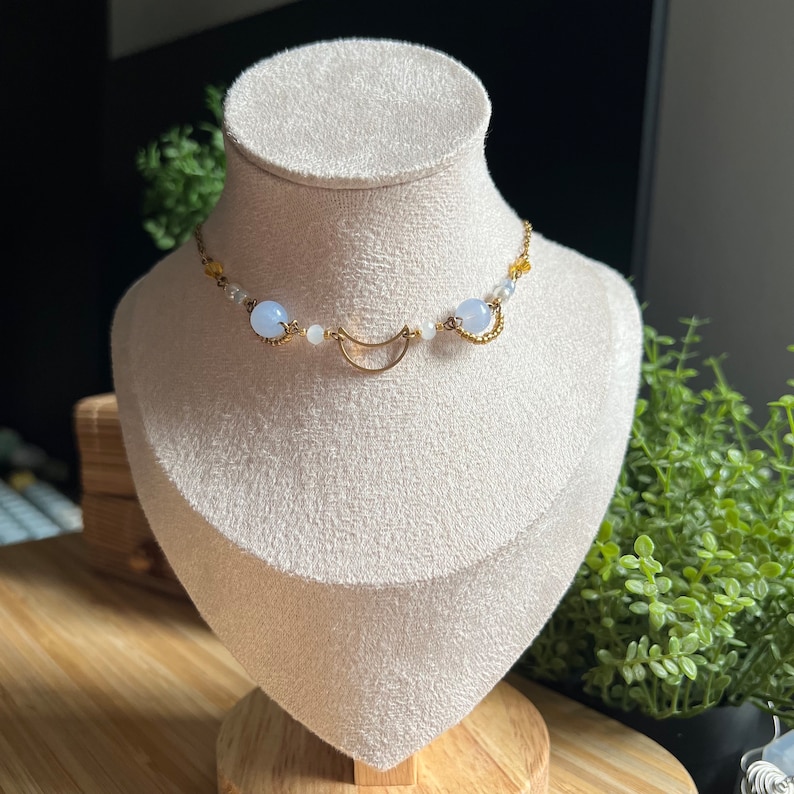 Op de afbeelding: Een gouden ketting met een halve maan hanger, blauwe en witte kralen en gouden kettingschakels.