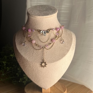 May include: A delicate gold layered necklace with pink beads and a crystal butterfly charm. The necklace has a dangling sun charm at the bottom.
