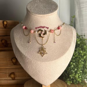 May include: A delicate necklace with pink and white beads, gold chains, and a gold pendant featuring a crescent moon and star design. The text "@asillyfrog" is visible on the necklace.