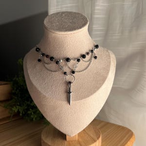 May include: A silver chain choker necklace with black beads and a silver dagger pendant. The necklace is layered with multiple chains and has a delicate, gothic aesthetic.