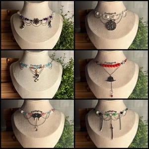 May include: A collection of six silver-toned chain chokers displayed on mannequin necks. Each necklace features unique charms, including floral, fruit, hat, and sword motifs, accented with colourful beads and layered silver chains.