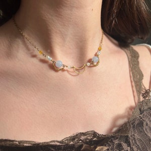 Op de afbeelding: Een gouden halve maan hanger ketting met lichtblauwe en witte kralen. De ketting wordt gedragen door een model dat een bruine kant top draagt.