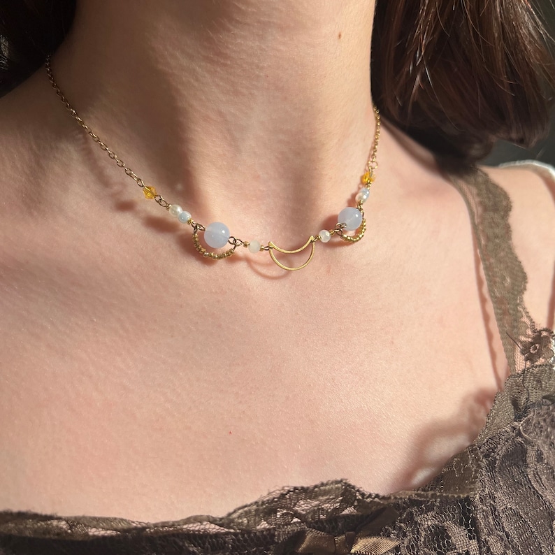 Op de afbeelding: Een gouden halve maan hanger ketting met lichtblauwe en witte kralen. De ketting wordt gedragen door een model dat een bruine kant top draagt.