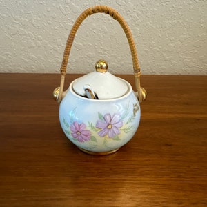 May include: A white ceramic sugar bowl with a floral design and a woven handle. The bowl has a lid with a small spoon inside. The bowl is decorated with purple and pink flowers on a light blue background.