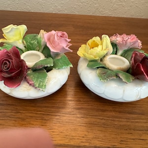 Op de afbeelding: Twee witte porseleinen kaarsenhouders met bloemmotieven. Elke houder heeft een ring van rozen, anjers en bladeren rond een kleine kaarsbeker.