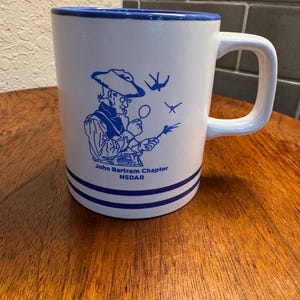 May include: White ceramic mug with a blue rim, handle, and decorative stripes. The mug features a blue illustration of a man with a magnifying glass and the text "John Bartram Chapter NSDAR".