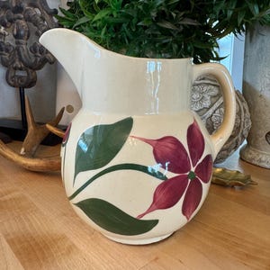 May include: A vintage cream-colored pitcher with a floral design. The pitcher has a wide mouth and a handle. The flowers are painted in shades of pink and green.