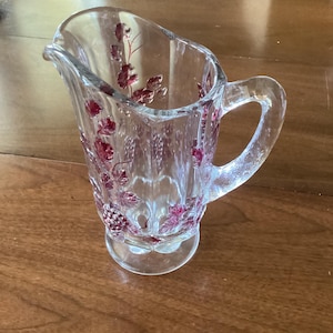 May include: Clear glass pitcher with a handle and a spout. The pitcher is decorated with a red floral pattern. The pitcher has a round base and a clear handle. The pitcher is likely used for serving beverages.