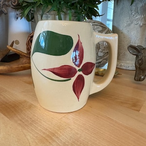 May include: A cream-coloured ceramic mug with a red and green floral design. The mug has a rounded body and a handle.