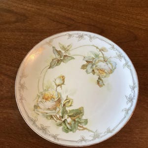 May include: A round, white porcelain plate with a gold rim. The plate features a hand-painted design of yellow roses with green leaves and buds. A delicate floral border encircles the edge of the plate, adding to its vintage charm.