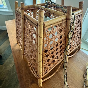 May include: A vintage, square-shaped rattan pendant light fixture. The light has a woven cane design on the sides and a light-colored frame. A metal chain and a light bulb are visible inside the fixture.