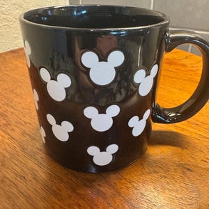 May include: Black ceramic mug with white Mickey Mouse head silhouettes. The mug has a glossy finish and a large handle. The mug is sitting on a wooden surface.