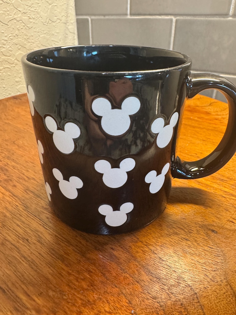 May include: Black ceramic mug with white Mickey Mouse head silhouettes. The mug has a glossy finish and a large handle. The mug is sitting on a wooden surface.