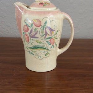 Puede incluir: Una jarra de crema de cerámica vintage con tapa rosa y diseño floral. La jarra está decorada con flores y hojas rosas, moradas y verdes.