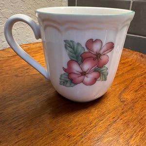 Puede incluir: Taza de cerámica blanca con asa curva, con un diseño floral de flores rosas y hojas verdes. El borde está adornado con un borde verde claro. La taza está sobre una superficie de madera.