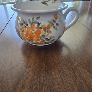 Puede incluir: Un macetero de cerámica blanca con un diseño floral con flores naranjas, amarillas y marrones. El macetero tiene una base ancha y redonda y un asa en un lado.