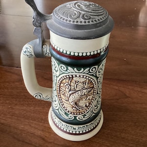 Pode incluir: Um caneco de cerveja de cerâmica com tampa de metal articulada. O caneco apresenta uma base creme, uma faixa decorativa verde e castanha e uma imagem central de um peixe. A pega e a tampa são de metal. A base diz "the strike rainbow trout".