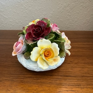 May include: A porcelain flower arrangement featuring a bouquet of roses in shades of pink, red, yellow, and orange. The roses are arranged in a white ceramic vase with a textured surface.