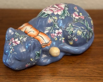 May include: A blue porcelain cat figurine with floral designs in pink, yellow, and green. The cat is lying down with its head resting on its paws.