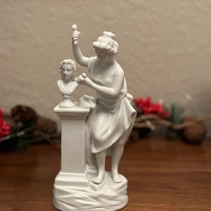 May include: White porcelain sculpture of a woman holding a chisel and working on a bust of a person's head. The sculpture is on a pedestal with a base.