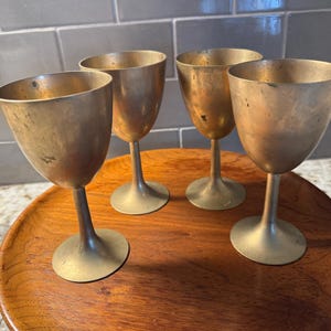 Pode incluir: Quatro cálices de latão vintage com uma taça em forma de tulipa e haste fina, sobre uma bandeja de madeira. Os cálices têm uma aparência envelhecida e manchada, sugerindo que são antigos ou vintage. O latão tem um tom dourado quente.