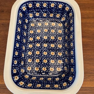 Puede incluir: Un plato para hornear de cerámica blanca con un patrón geométrico azul y amarillo. El plato tiene una forma rectangular y un borde ligeramente elevado.