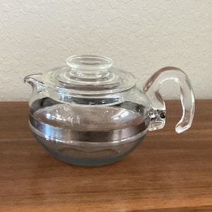 May include: A clear glass teapot with a silver band around the middle. The teapot has a lid and a handle.
