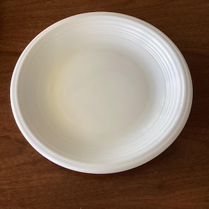 Puede incluir: Un plato de cerámica blanco con un centro ligeramente hundido y una serie de crestas concéntricas alrededor del borde. El plato es redondo y parece estar limpio. El plato está sobre una superficie de madera.